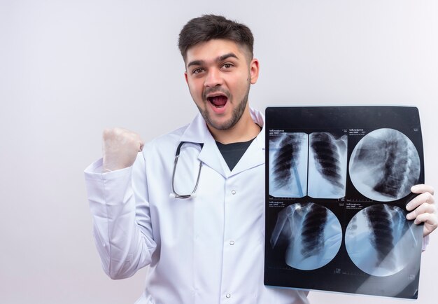 Joven médico guapo con bata médica blanca, guantes médicos blancos y un estetoscopio feliz con los resultados de la tomografía de pie sobre la pared blanca