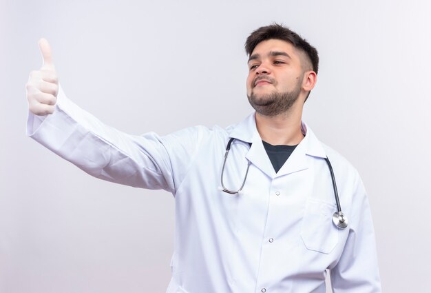 Joven médico guapo con bata médica blanca, guantes médicos blancos y un estetoscopio haciendo felices pulgares hacia arriba mirando además de estar parado sobre la pared blanca