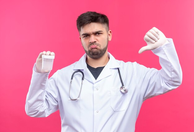 Joven médico guapo con bata médica blanca guantes médicos blancos y un estetoscopio haciendo infelices pulgares hacia abajo para obtener resultados en un contenedor de análisis transparente de pie sobre fondo rosa