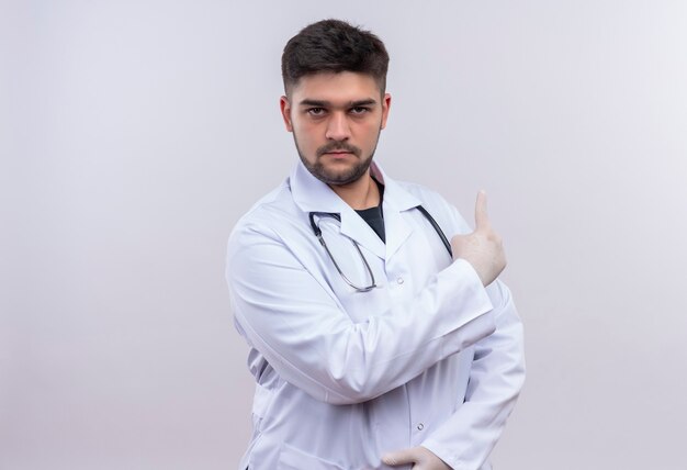 Joven médico guapo con bata médica blanca, guantes médicos blancos y un estetoscopio mirando seriamente mostrando la espalda con el dedo índice de pie sobre la pared blanca
