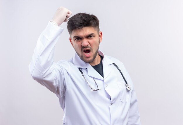 Joven médico guapo con bata médica blanca, guantes médicos blancos y un estetoscopio que amenaza con el puño parado sobre la pared blanca