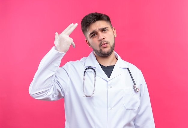 Joven médico guapo con bata médica blanca, guantes médicos blancos y un estetoscopio que muestra una cara fría haciendo pistola con los dedos de pie sobre la pared rosada