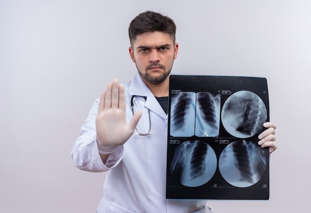 Joven médico guapo con bata médica blanca, guantes médicos blancos y un estetoscopio que muestra la señal de stop con tomografía de pie sobre una pared blanca