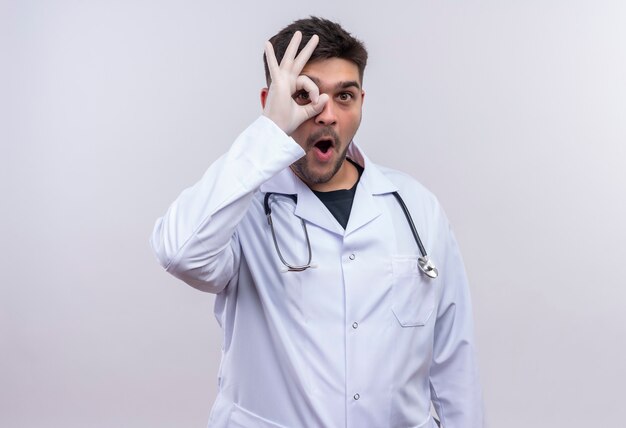 Joven médico guapo con bata médica blanca, guantes médicos blancos y un estetoscopio que muestra el signo de ok con la mano sobre la pared blanca