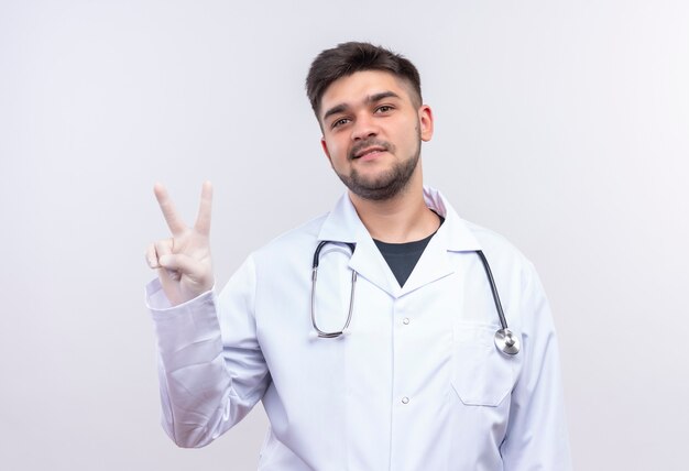 Joven médico guapo con bata médica blanca, guantes médicos blancos y estetoscopio sonriendo mostrando el signo de la paz con los dedos de pie sobre la pared blanca