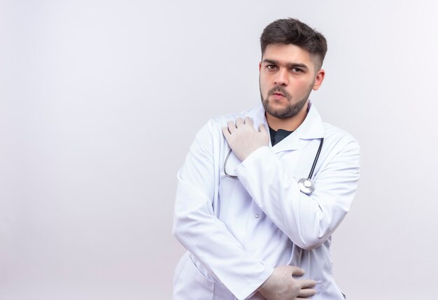 Joven médico guapo con bata médica blanca, guantes médicos blancos y estetoscopio sosteniendo el estómago y el hombro