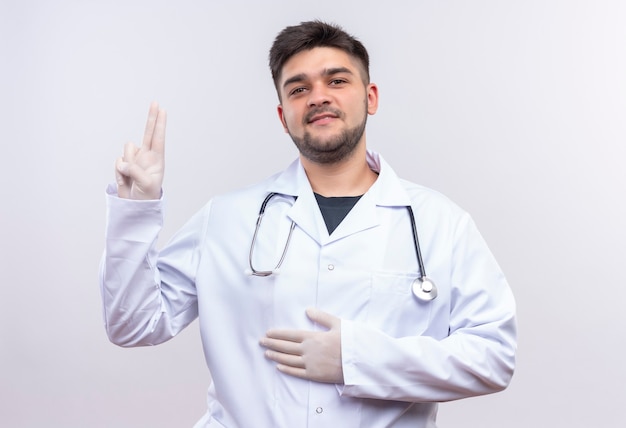 Joven médico guapo con bata médica blanca, guantes médicos blancos y estetoscopio sosteniendo el estómago mostrando el signo de la paz de pie sobre la pared blanca