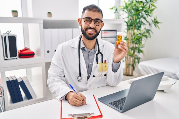 Joven médico hispano sosteniendo una botella de pastillas escribiendo en un documento en la clínica