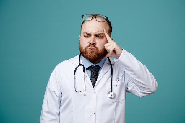 Joven médico inseguro con abrigo médico y estetoscopio alrededor del cuello y anteojos en la cabeza mirando al lado con un ojo cerrado mostrando un gesto de pensamiento aislado en el fondo azul