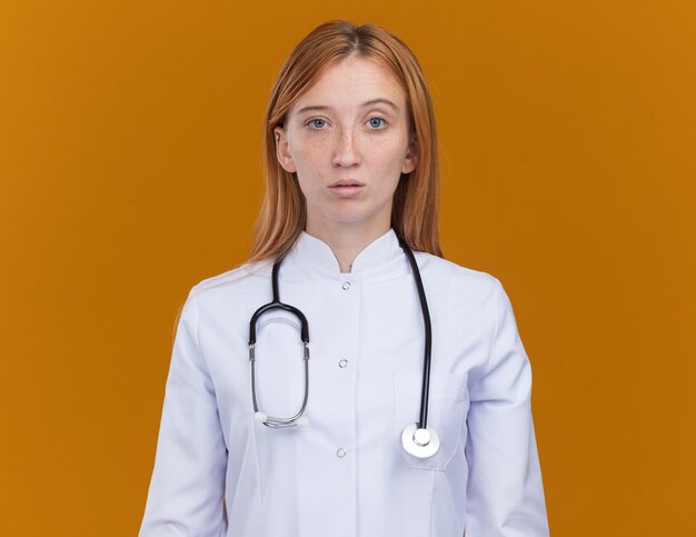 Joven médico de jengibre vistiendo una bata médica y un estetoscopio aislado en la pared naranja