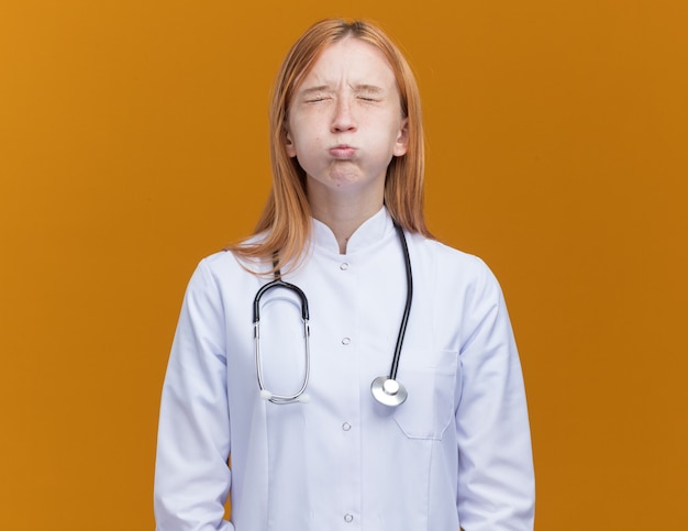 Joven médico de jengibre vistiendo bata médica y estetoscopio hinchando las mejillas con los ojos cerrados aislados en la pared naranja