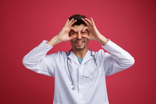 Joven médico juguetón con uniforme médico y estetoscopio alrededor del cuello mirando a la cámara mostrando un gesto de binoculares aislado en el fondo rojo