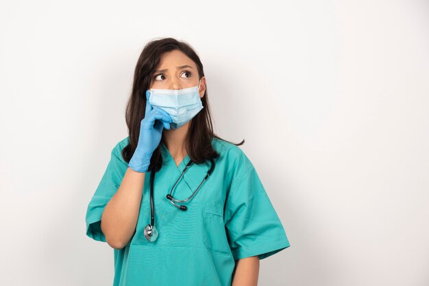 Joven médico en máscara médica y guantes pensando sobre fondo blanco. Foto de alta calidad