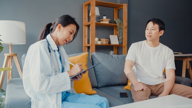 Joven médico médico profesional femenino de Asia usando tableta digital compartiendo buenas noticias sobre pruebas de salud con paciente masculino feliz sentarse en el sofá en casa