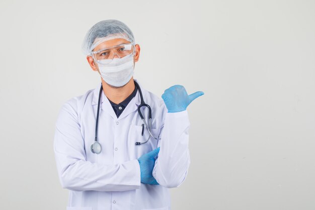 Joven médico mostrando con el pulgar hacia arriba en bata blanca, gorro y guantes