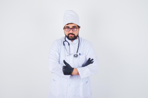 Joven médico mostrando el pulgar hacia arriba en uniforme blanco y mirando confiado, vista frontal.