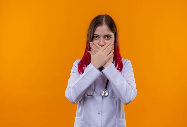 Joven médico mujer vistiendo estetoscopio bata médica boca cubierta con las manos en la pared naranja aislada