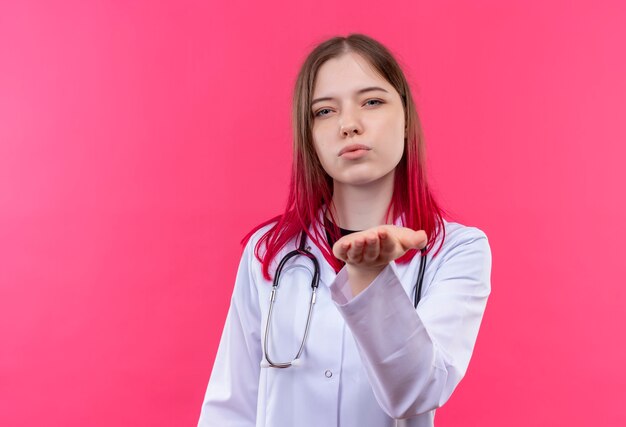 Joven médico mujer vistiendo estetoscopio bata médica mostrando gesto de beso en rosa pared aislada