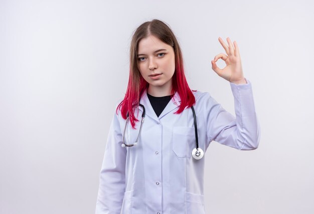 Joven médico mujer vistiendo estetoscopio bata médica mostrando gesto okey en pared blanca aislada