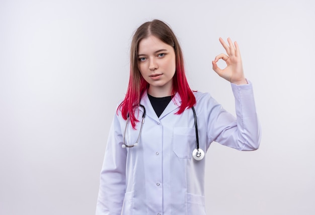 Joven médico mujer vistiendo estetoscopio bata médica mostrando gesto okey en pared blanca aislada