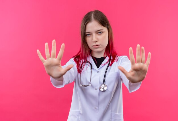 Joven médico mujer vistiendo un estetoscopio bata médica mostrando gesto de parada en rosa pared aislada