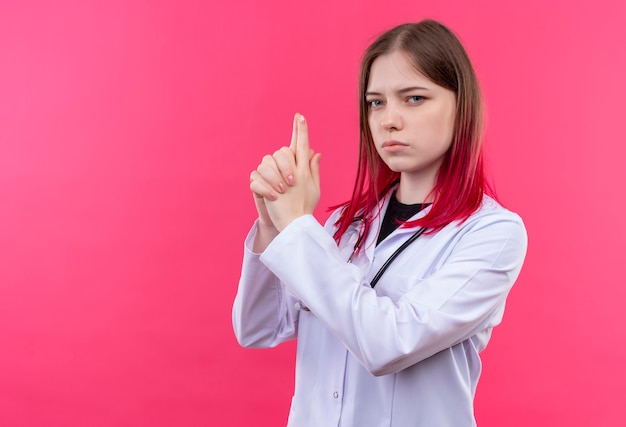 Joven médico mujer vistiendo estetoscopio bata médica mostrando gesto de pistola en rosa pared aislada