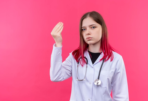 Joven médico mujer vistiendo estetoscopio bata médica mostrando gesto de punta en rosa pared aislada