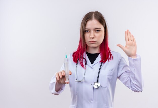 Joven médico mujer vistiendo estetoscopio bata médica sosteniendo una jeringa mostrando gesto de parada en la pared blanca aislada