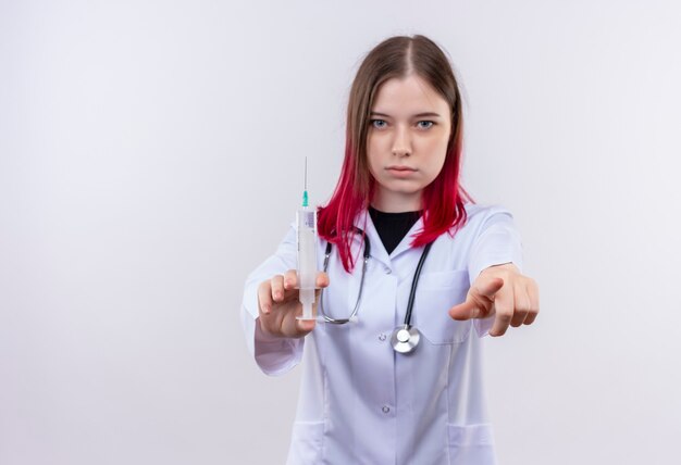 Joven médico mujer vistiendo estetoscopio bata médica sosteniendo una jeringa mostrando su gesto en la pared blanca aislada