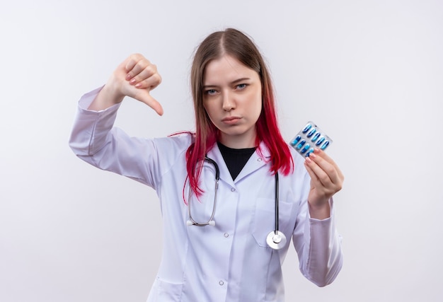 Joven médico mujer vistiendo estetoscopio bata médica sosteniendo pastillas con el pulgar hacia abajo en la pared blanca aislada