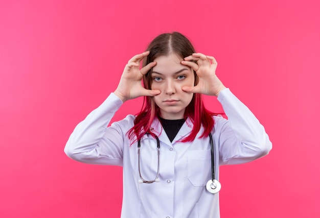 Joven médico mujer vistiendo estetoscopio bata médica sosteniendo sus ojos abiertos con las manos en la pared rosa aislada