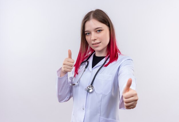 Joven médico mujer vistiendo estetoscopio bata médica sus pulgares hacia arriba en la pared blanca aislada