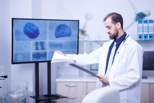 Joven médico pasando páginas con análisis en su portapapeles en un laboratorio para problemas cerebrales. Cerebro mostrado en el monitor.