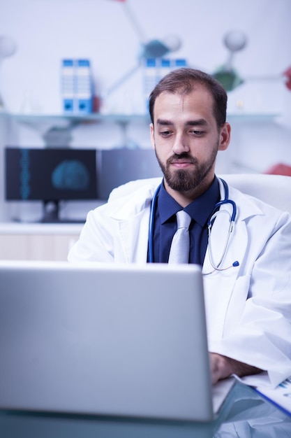 Joven médico pensando y trabajando en su laptop en un laboratorio privado. Expresión de doctor haciendo su trabajo.