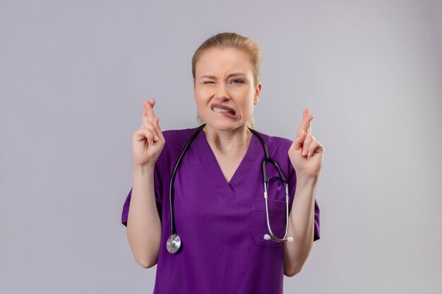 Joven médico preocupado vistiendo una bata médica púrpura y un estetoscopio cruzando los dedos en la pared blanca aislada