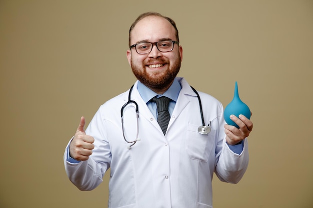 Un joven médico sonriente con bata de laboratorio y estetoscopio alrededor del cuello mirando a la cámara sosteniendo un enema mostrando el pulgar hacia arriba aislado en un fondo verde oliva