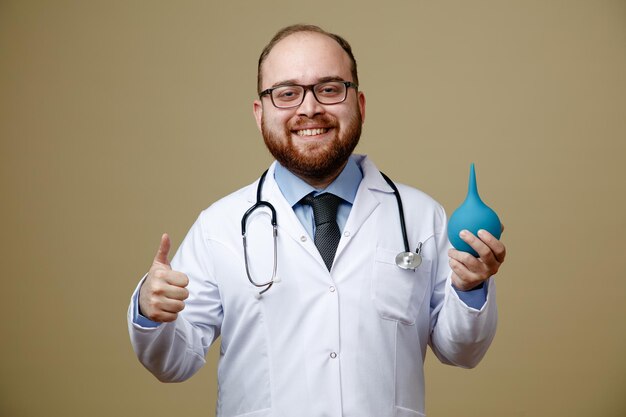 Un joven médico sonriente con bata de laboratorio y estetoscopio alrededor del cuello mirando a la cámara sosteniendo un enema mostrando el pulgar hacia arriba aislado en un fondo verde oliva