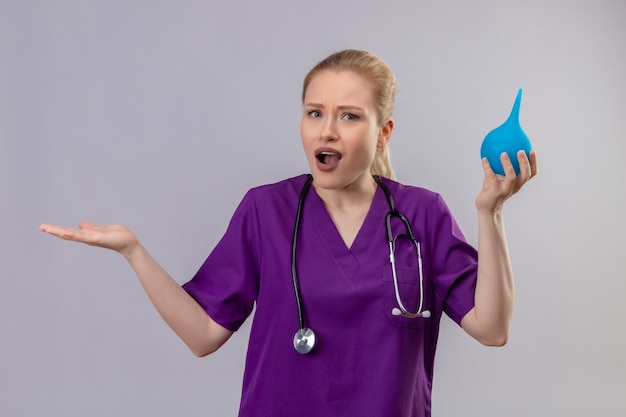 Joven médico sorprendido vistiendo una bata médica púrpura y un estetoscopio sosteniendo enema sobre fondo blanco aislado