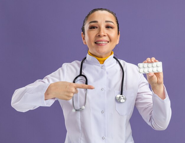 Joven médico sosteniendo blister con pastillas apuntando con el dedo índice sonriendo confiado