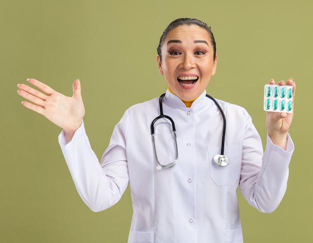 Joven médico sosteniendo blister con pastillas feliz y emocionado con el brazo levantado