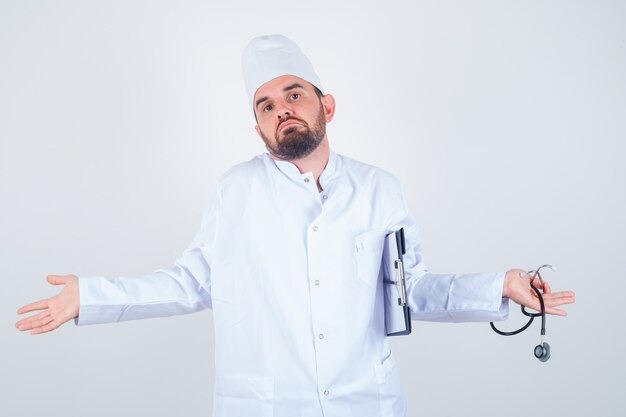 Joven médico sosteniendo el portapapeles y un estetoscopio, mostrando un gesto de impotencia encogiéndose de hombros en uniforme blanco y mirando perplejo. vista frontal.