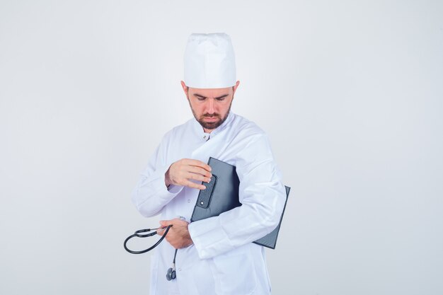 Joven médico sosteniendo el portapapeles y un estetoscopio en uniforme blanco y mirando pensativo, vista frontal.