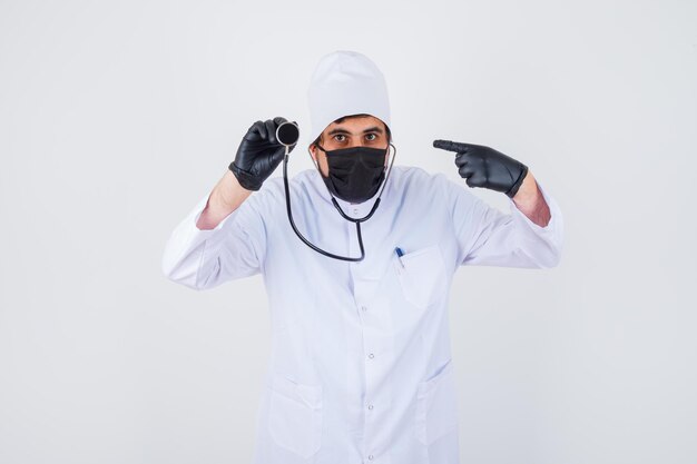 Joven médico en uniforme blanco apuntando con el estetoscopio y mirando confiado, vista frontal.