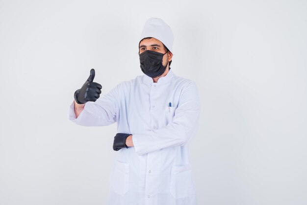 Joven médico en uniforme blanco mostrando el pulgar hacia arriba y mirando confiado, vista frontal.