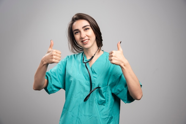 Joven médico en uniforme dando pulgar hacia arriba en la pared gris.