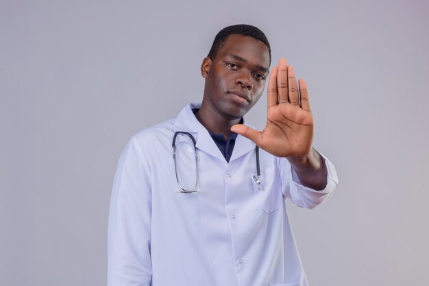 Joven médico varón afroamericano vistiendo bata blanca con estetoscopio con cara seria con la mano abierta haciendo señal de stop
