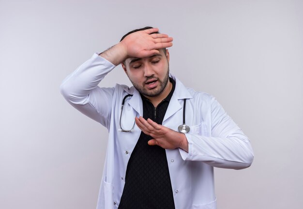 Joven médico varón barbudo con bata blanca con estetoscopio con aspecto cansado y aburrido con dolor de cabeza