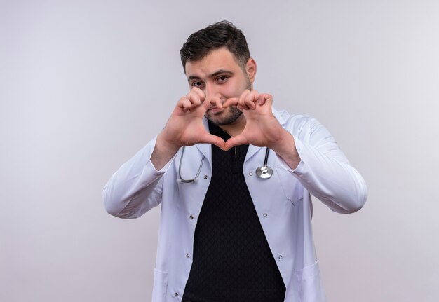 Joven médico varón barbudo con bata blanca con estetoscopio haciendo gesto de corazón con los dedos
