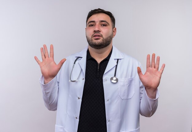 Joven médico varón barbudo con bata blanca con estetoscopio levantando los brazos en señal de rendición con expresión de miedo