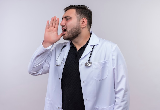 Joven médico varón barbudo con bata blanca con estetoscopio llamando a alguien con la mano cerca de la boca